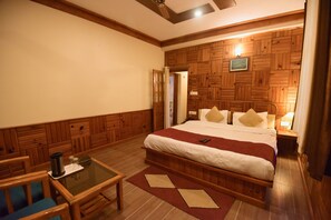 A hotel room with a wooden paneled wall, a large bed with pillows, a bedside table with a lamp, a small table with a coffee pot, and a chair.