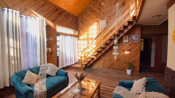 A cozy room with wooden walls and a sloped ceiling, featuring two teal sofas with macrame pillows, a wooden coffee table, and a staircase with wooden railings. - Boho Mansion - by Dumnu Homes (Manali)