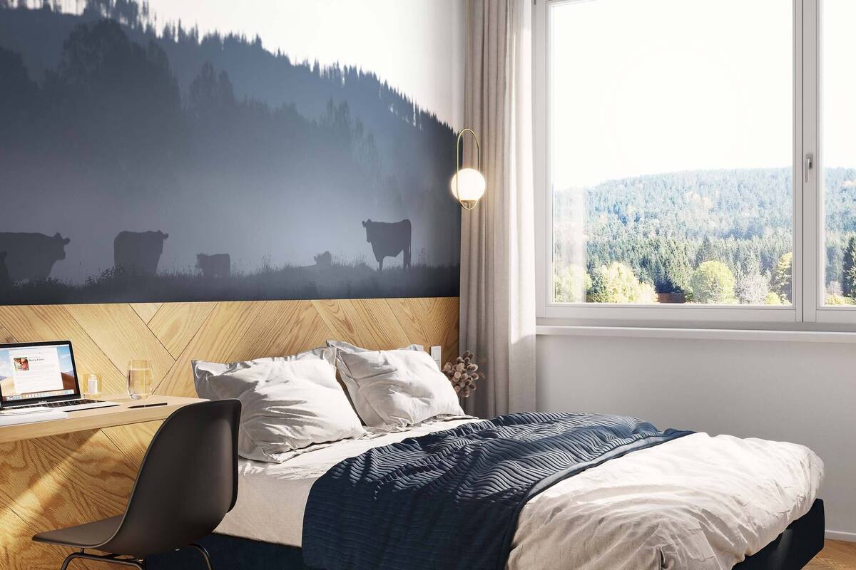Un dormitorio con cama, escritorio con ordenador portátil, silla y ventana con vista al bosque.