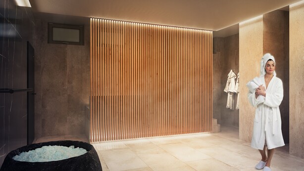 Ein Wellnessbereich mit einer Wand aus Holzpaneelen, einer Badewanne aus schwarzem Marmor und einer Person im Bademantel.