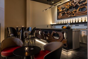 Zona de bar moderna con una escultura metálica única, sillas negras y rojas, y una vitrina para vinos.