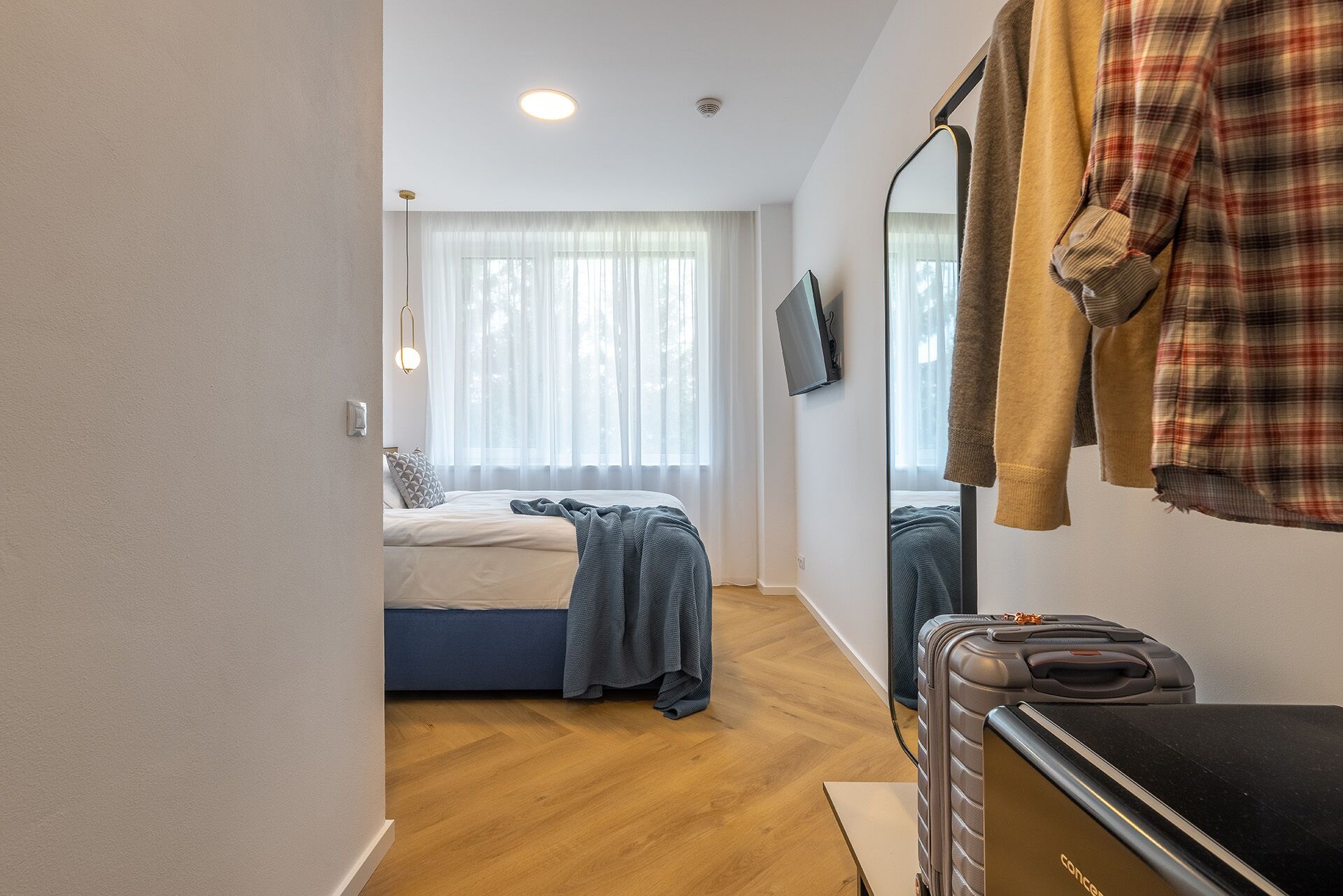Ein Schlafzimmer mit einem Bett, einem Koffer, einem Spiegel und einem Fernseher an der Wand.