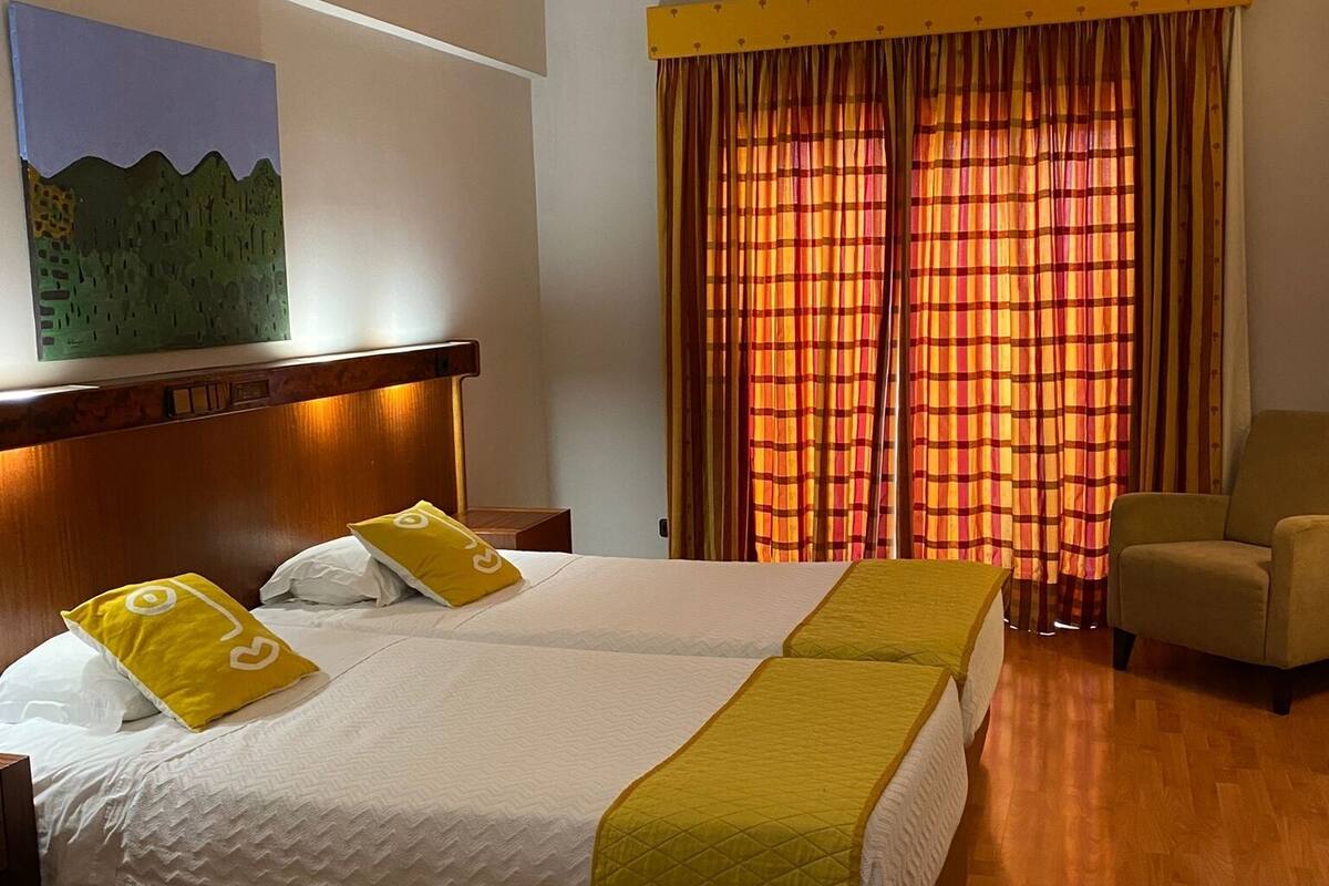 A hotel room with a double bed, two pillows, a chair, a painting on the wall, and a window with curtains.