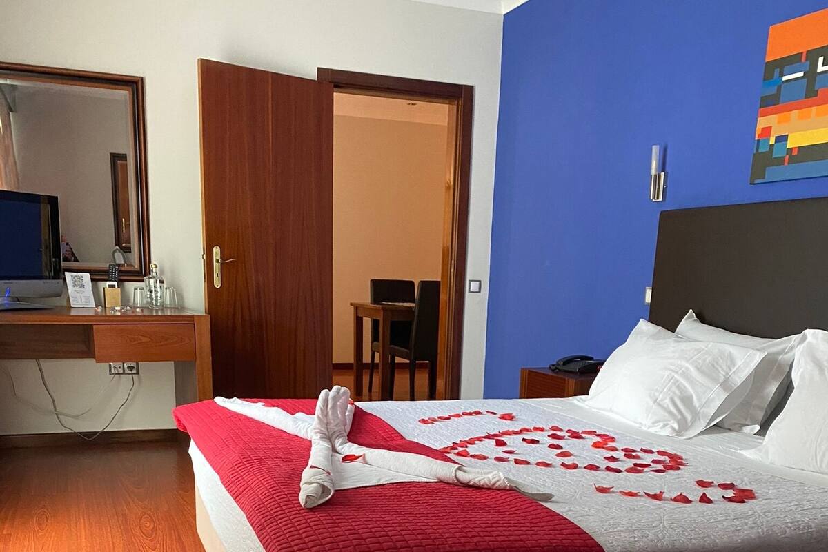A hotel room with a bed, a desk with a computer, a chair, and a door leading to another room.