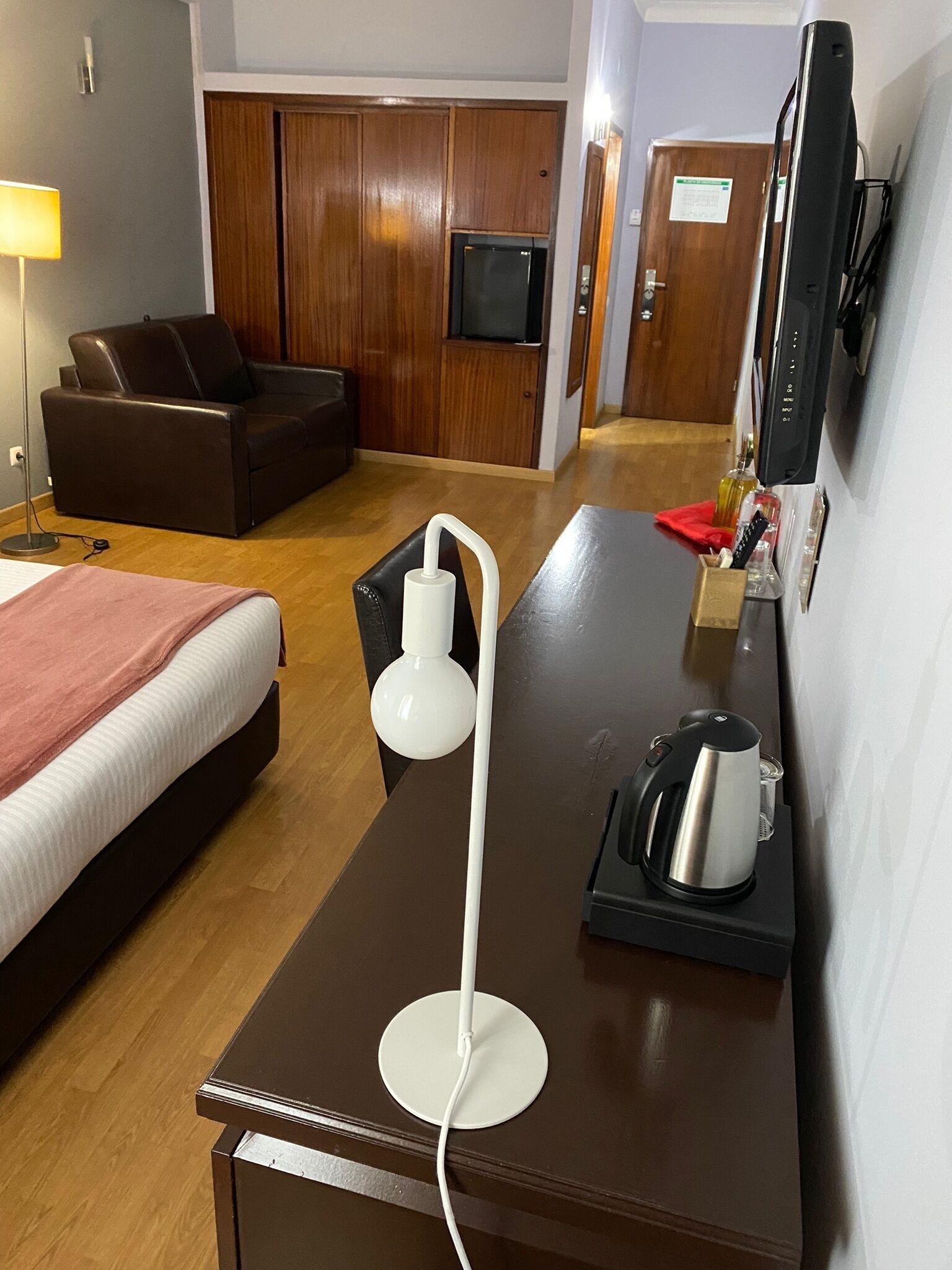 A hotel room with a bed, a desk with a coffee maker, a lamp, and a TV mounted on the wall.