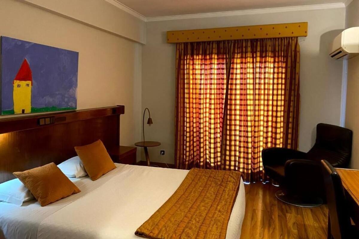 A hotel room with a bed, a desk, and a chair. There is a painting on the wall and a window with curtains.