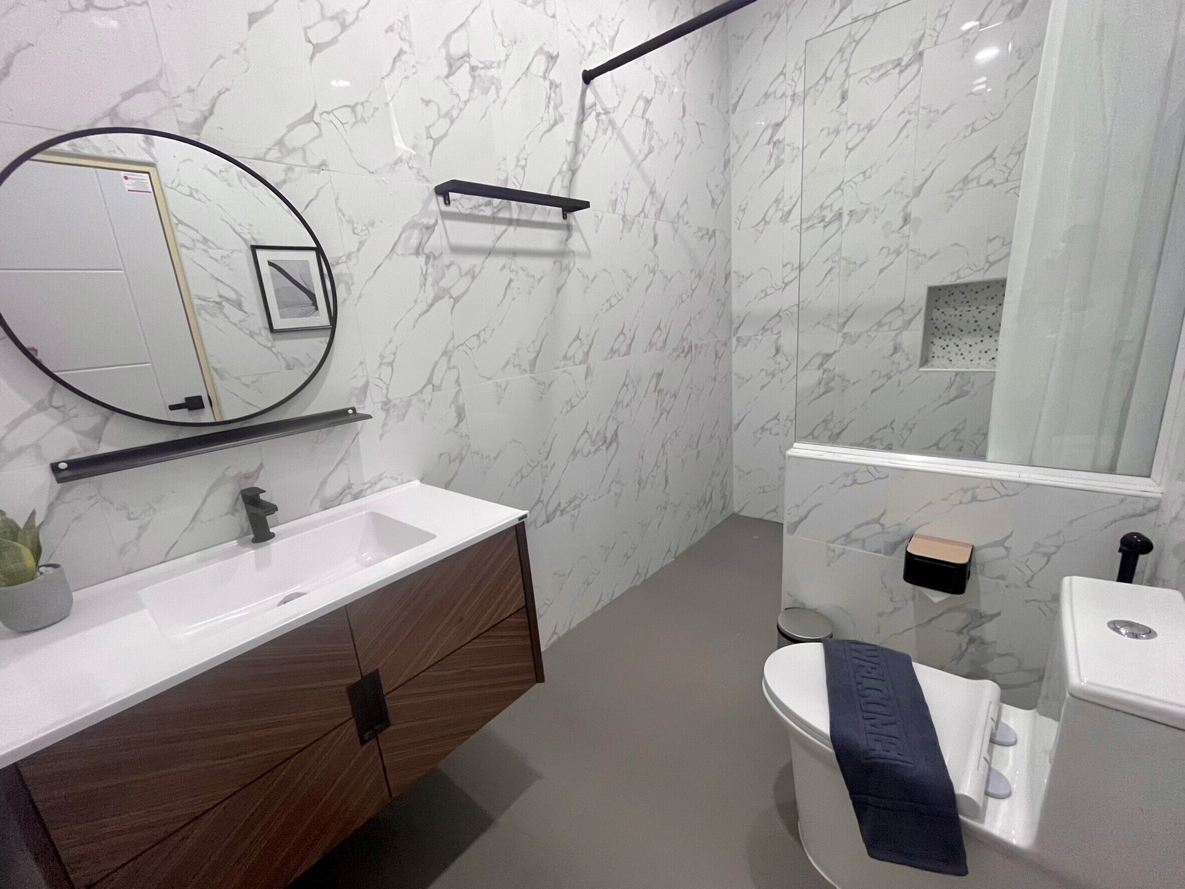 Un baño moderno con salpicadero de mármol, tocador de madera, espejo redondo, inodoro y zona de ducha.