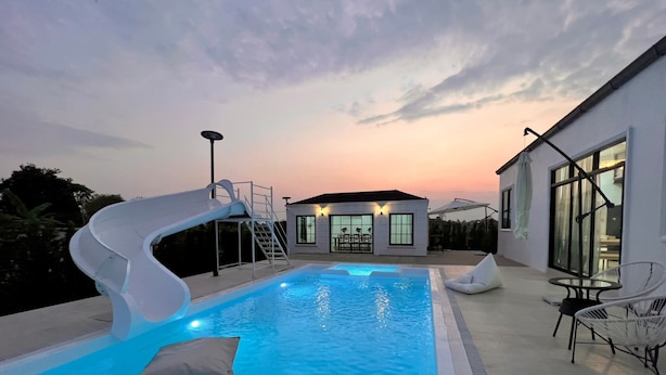 A swimming pool with a slide, a covered seating area, and a building with a circular window in the background.