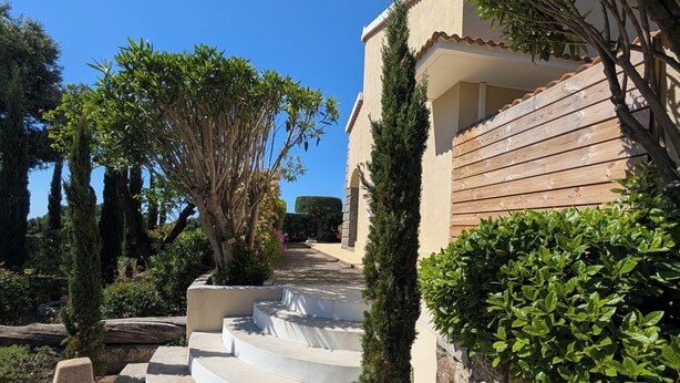 Une maison avec un escalier en pierre menant à une entrée fermée par un portail, entourée de verdure et sous un ciel bleu azur.