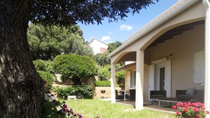 Un espace extérieur aménagé, abrité et doté de tables et de chaises, entouré d’un jardin avec des plantes en fleurs et un grand arbre.