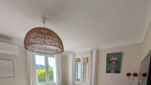 Une pièce comprenant une table en verre, des chaises, un canapé, une fenêtre avec des rideaux, un ventilateur de plafond et un meuble contenant des objets décoratifs.
