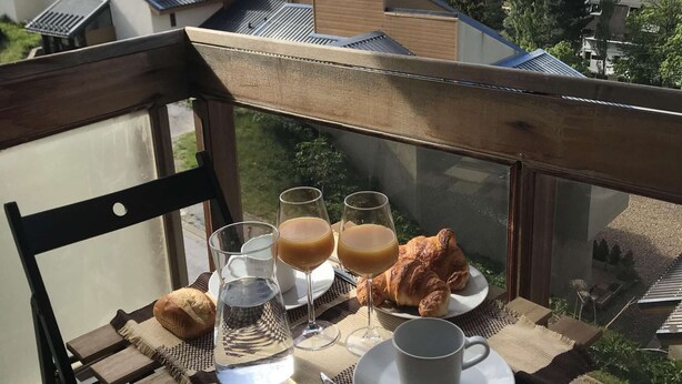 A balcony with a table set for breakfast, including croissants, glasses of juice, and a cup, overlooking a mountainous landscape. - Brand new "jungle" studio - panoramic view - parking - city center (Font-Romeu-Odeillo-Via)