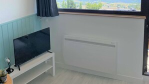 A room with a large window, a flat-screen TV on a white stand, and a radiator below the window. - Brand new "jungle" studio - panoramic view - parking - city center (Font-Romeu-Odeillo-Via)