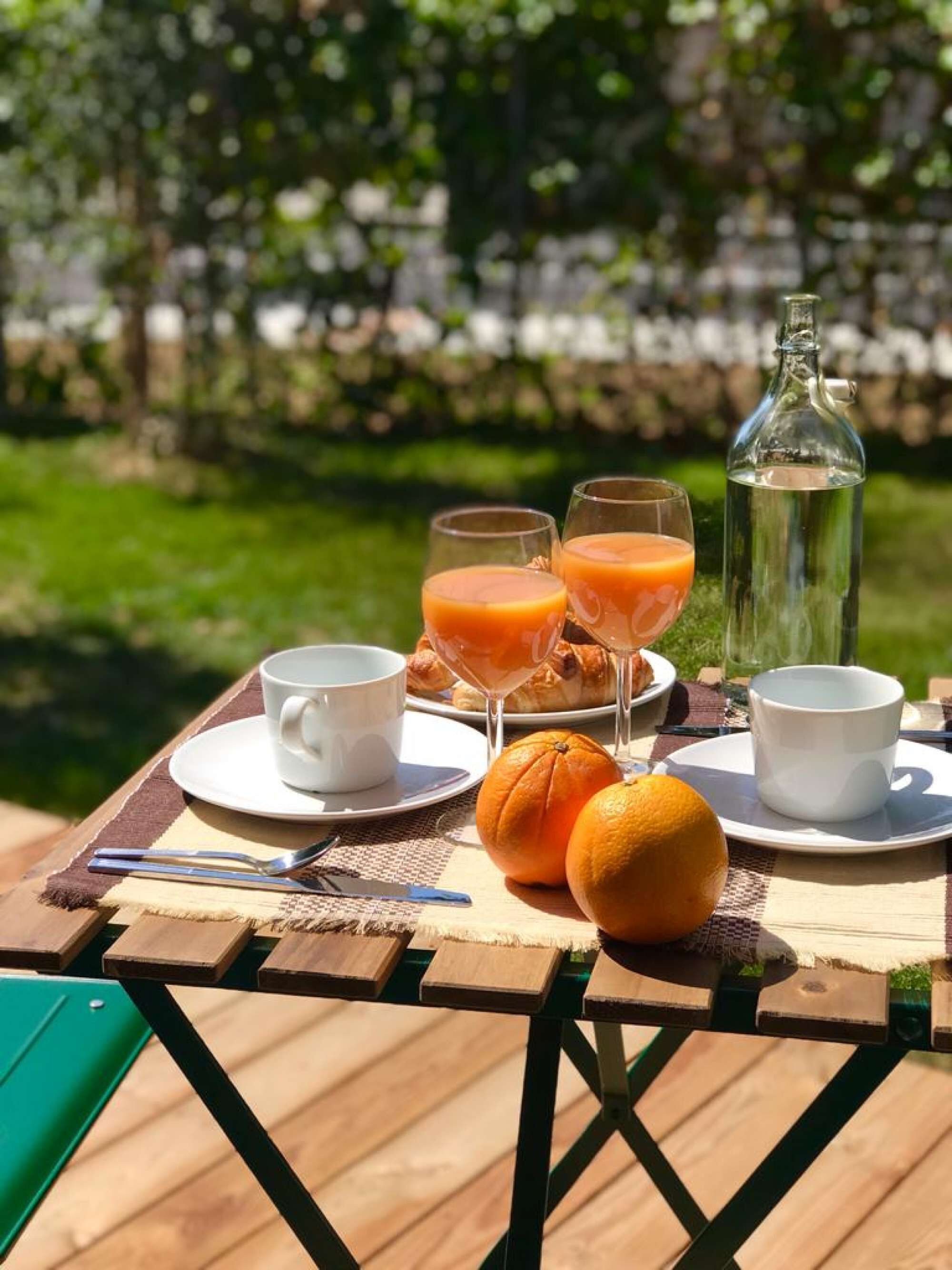 Ein Holztisch mit Frühstücksangeboten, darunter Croissants, Orangensaft und eine Wasserflasche, draußen mit einem grünen Stuhl.
