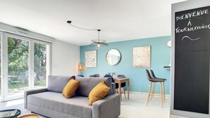 Uma sala de estar moderna com um sofá cinzento, duas mesinhas redondas, uma mesa de jantar com cadeiras, um espelho grande e uma lousa com a inscrição 'Bienvenue A Tournefeuille'.