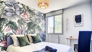 Quarto com cama, almofadas, escrivaninha, cadeira, janela e papel de parede tropical.