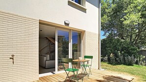Um deck de madeira com mesa e cadeiras, uma porta de correr de vidro que dá para um espaço interior, e uma parede externa de tijolos.
