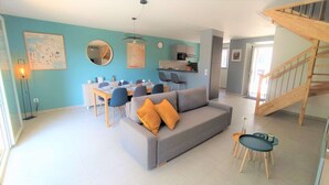 Uma sala de estar moderna com um sofá cinzento, duas poltronas azuis, uma mesa de jantar posta para quatro pessoas, uma área de cozinha e uma escada de madeira.