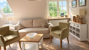 A cozy living room with a sofa, two armchairs, a wooden coffee table, and a white cabinet with drawers. There is a large window with a view of greenery outside.