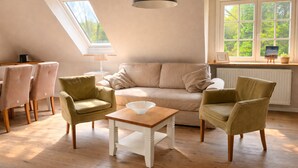 A cozy living room with a high ceiling, a skylight, a large window, a sofa, two armchairs, a small table, and a radiator.
