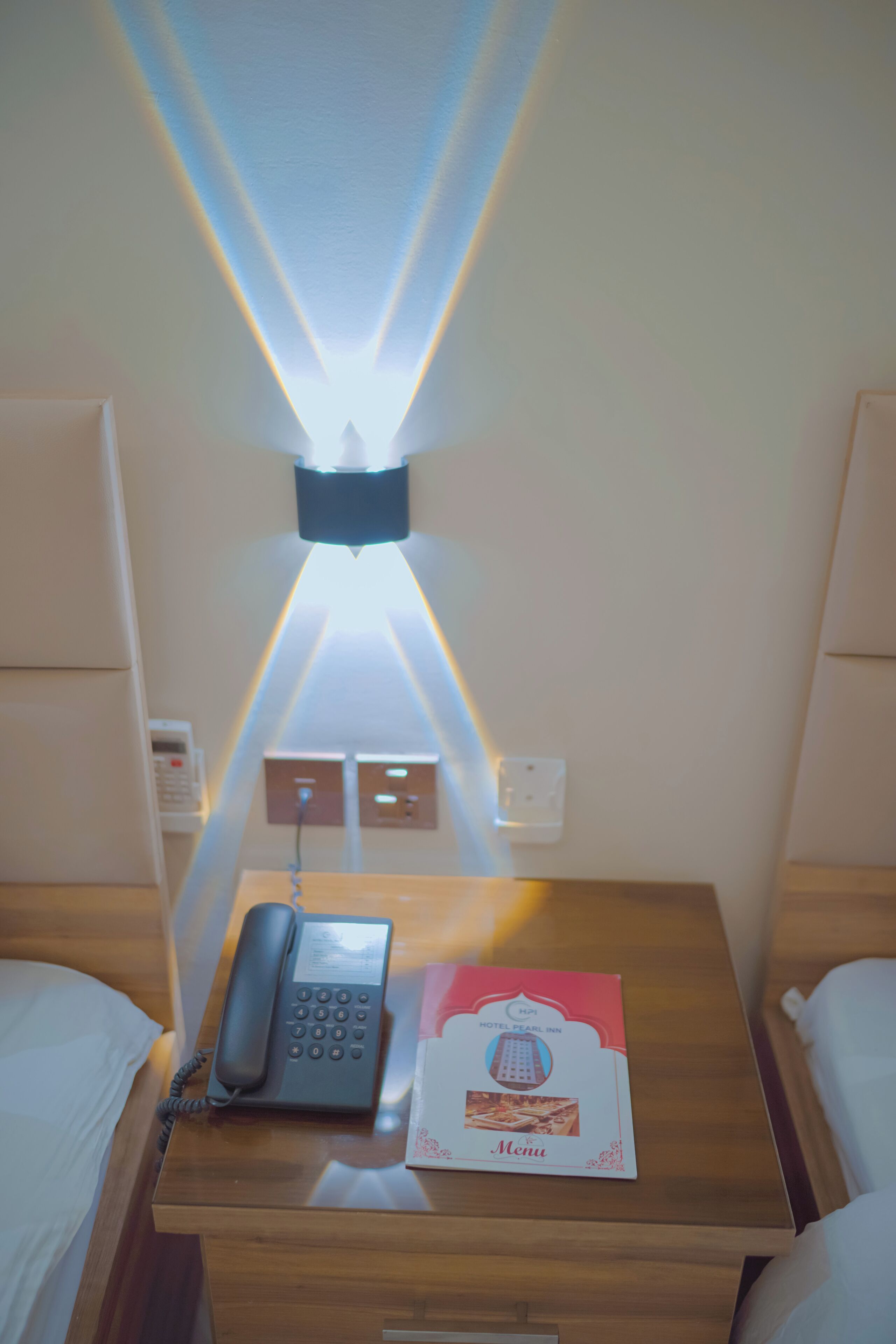 Habitación de hotel con una mesita de noche de madera, un teléfono, un menú y una lámpara fijada a la pared.