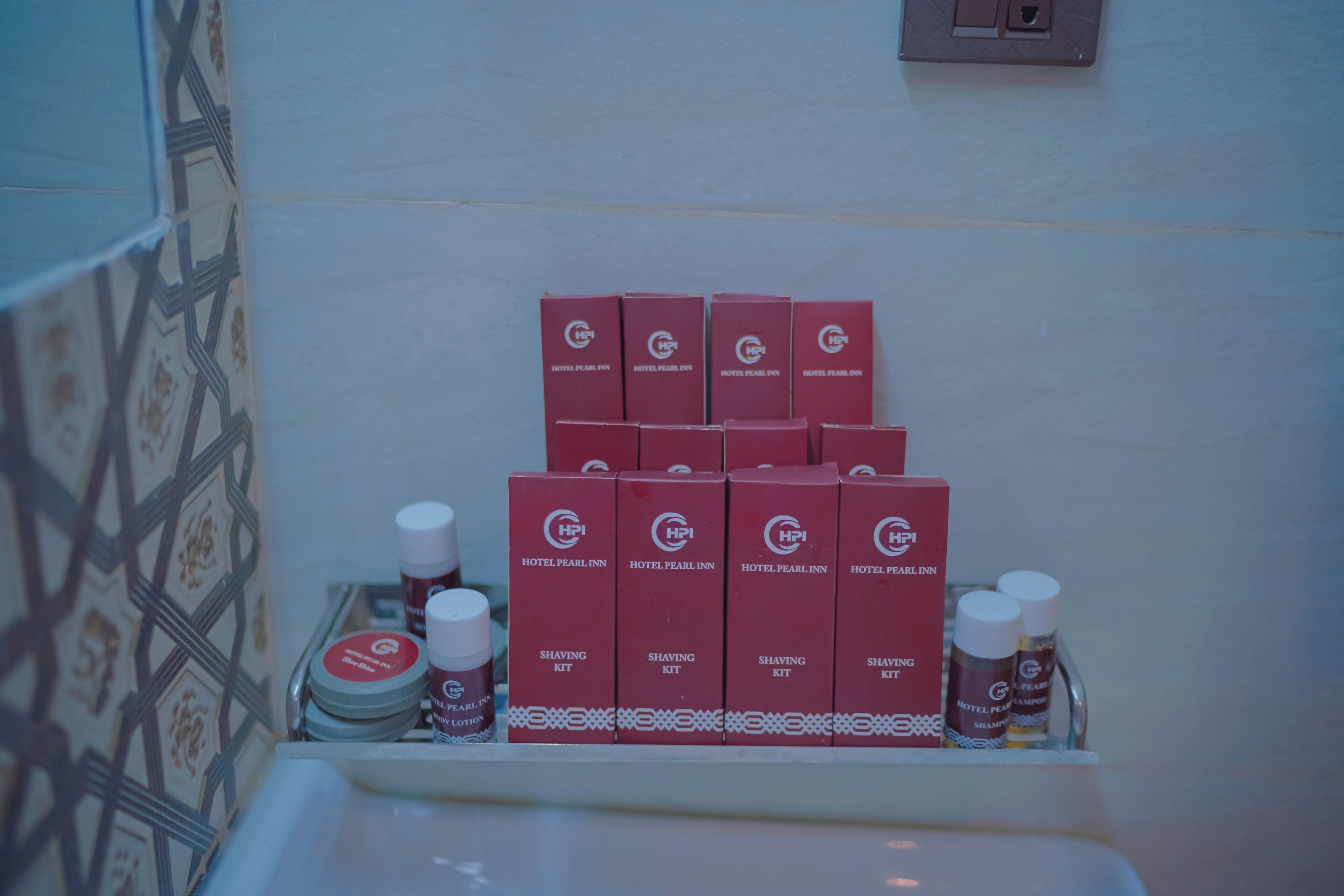 A bathroom with a tiled wall and a shelf holding several red boxes labeled 'Hotel Pearl Inn Shaving Kit' and bottles of shaving cream.