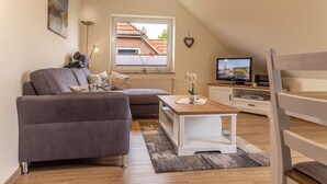 Moderni olohuone, jossa on harmaa sohva, puinen sohvapöytä, televisio ja ikkuna, josta on näkymä rakennukseen.