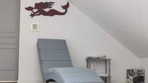 A room with a grey wall, a white ceiling, a grey floor, a grey chair with a blue cushion, a small table with a lamp, and a framed picture on the wall.