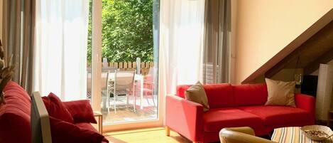 A living room with a red sofa, a yellow armchair, and a dining table with a bowl. There is a sliding glass door leading to an outdoor area with a table and chairs.