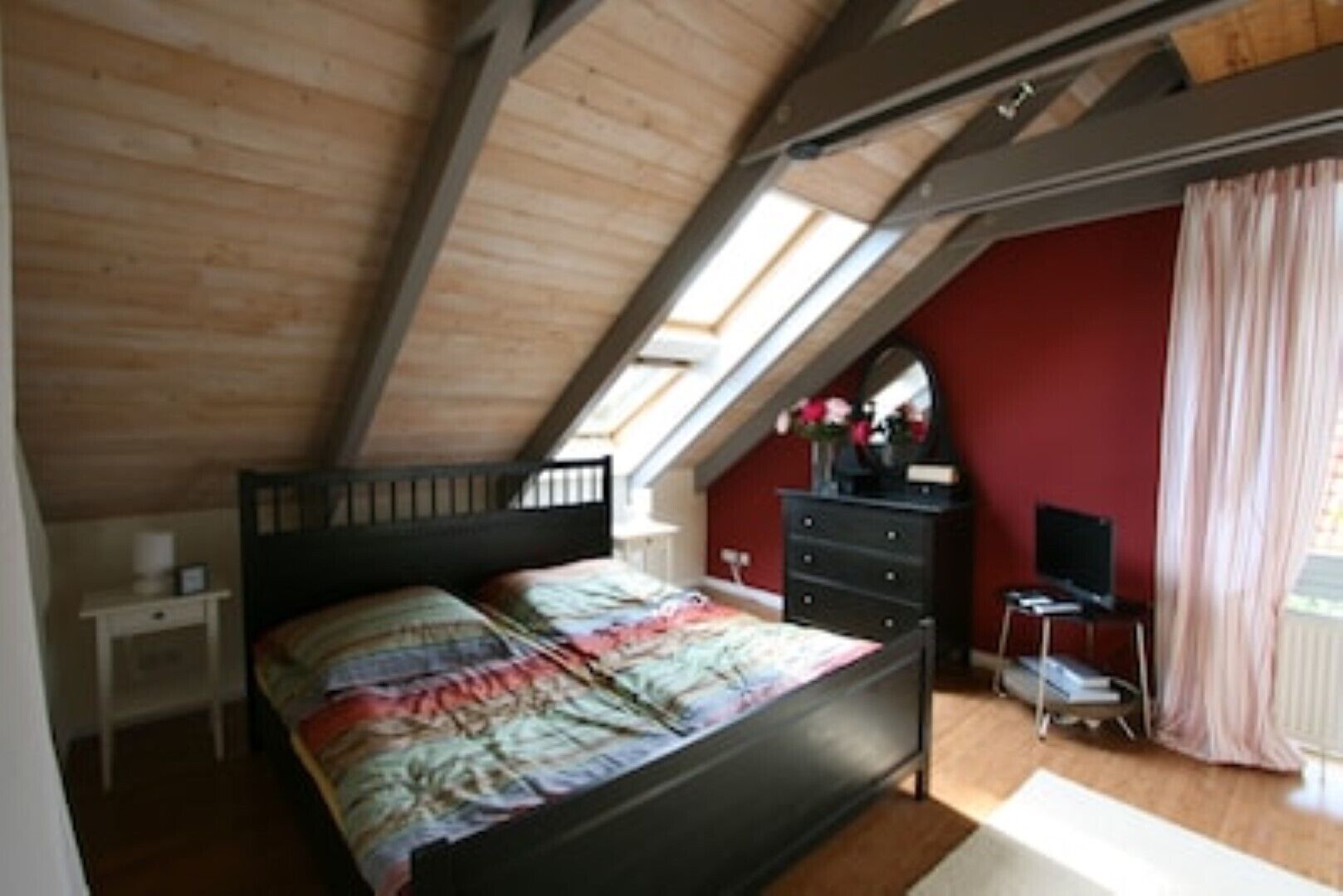 Ein Schlafzimmer mit schrägem Dach, einem Bett, einer Kommode, einem Fernseher und einem Fenster mit Vorhängen.
