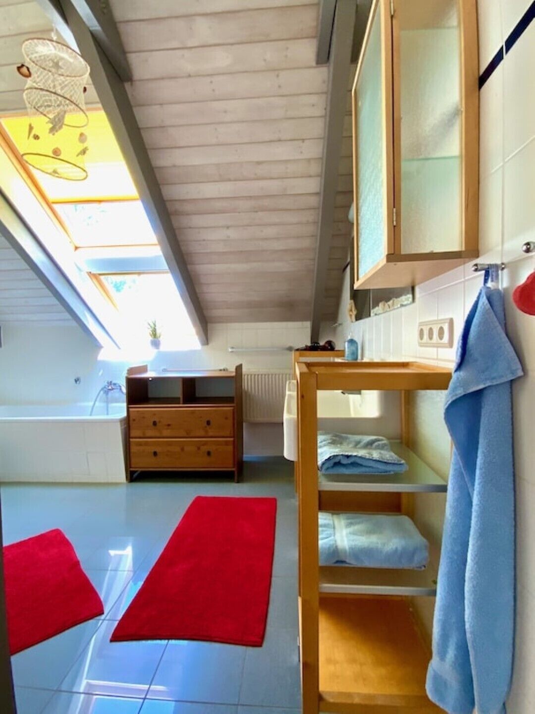 A room with a wooden ceiling, a window with a view, a wooden dresser, a red rug, and a blue towel on a shelf.