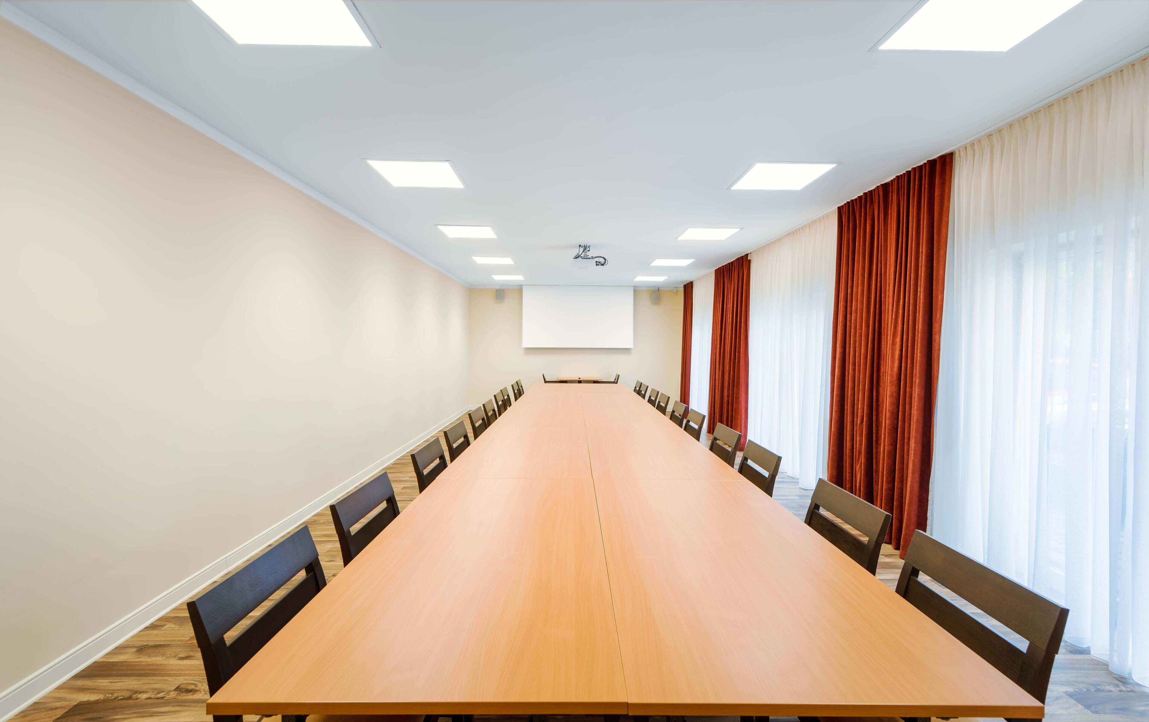 Ein Konferenzraum mit langem Tisch, Stühlen, einer Projektionsleinwand und Vorhängen.