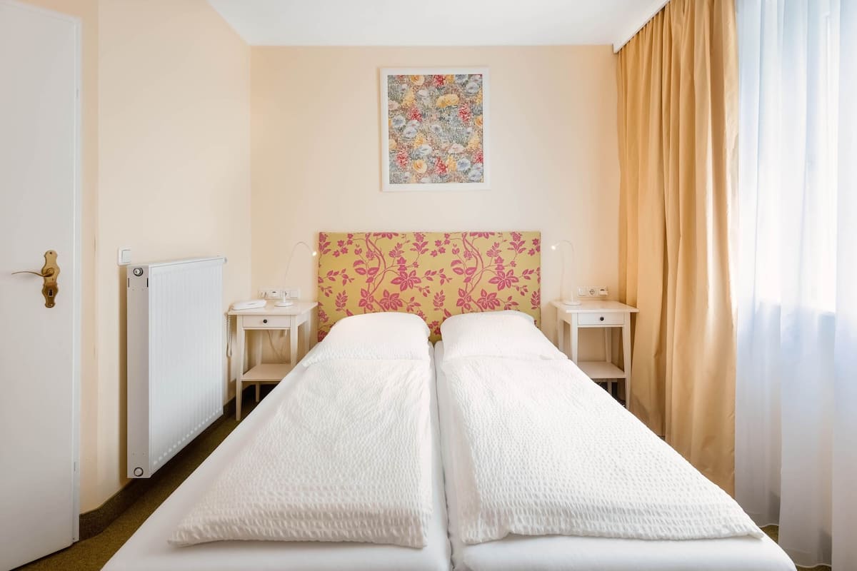 Habitación de hotel con dos camas individuales, un radiador, una mesita de noche con teléfono y un cabecero estampado con motivos florales.