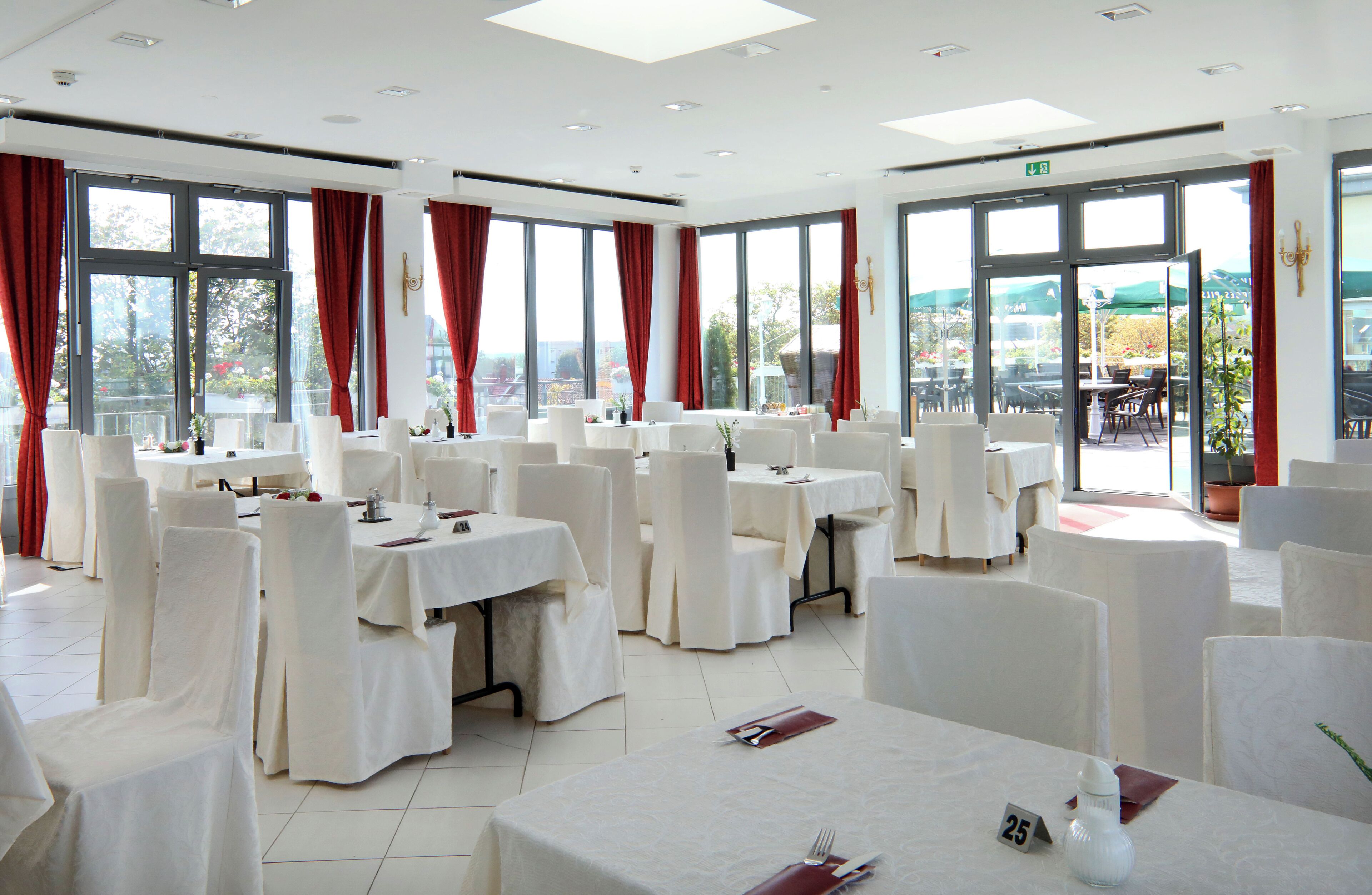A hotel dining area with multiple tables and chairs, large windows with red curtains, and a view of the outdoors.