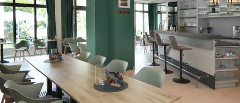 A modern dining area with a long wooden table, several chairs, and a bar area with shelves of glasses and bottles.