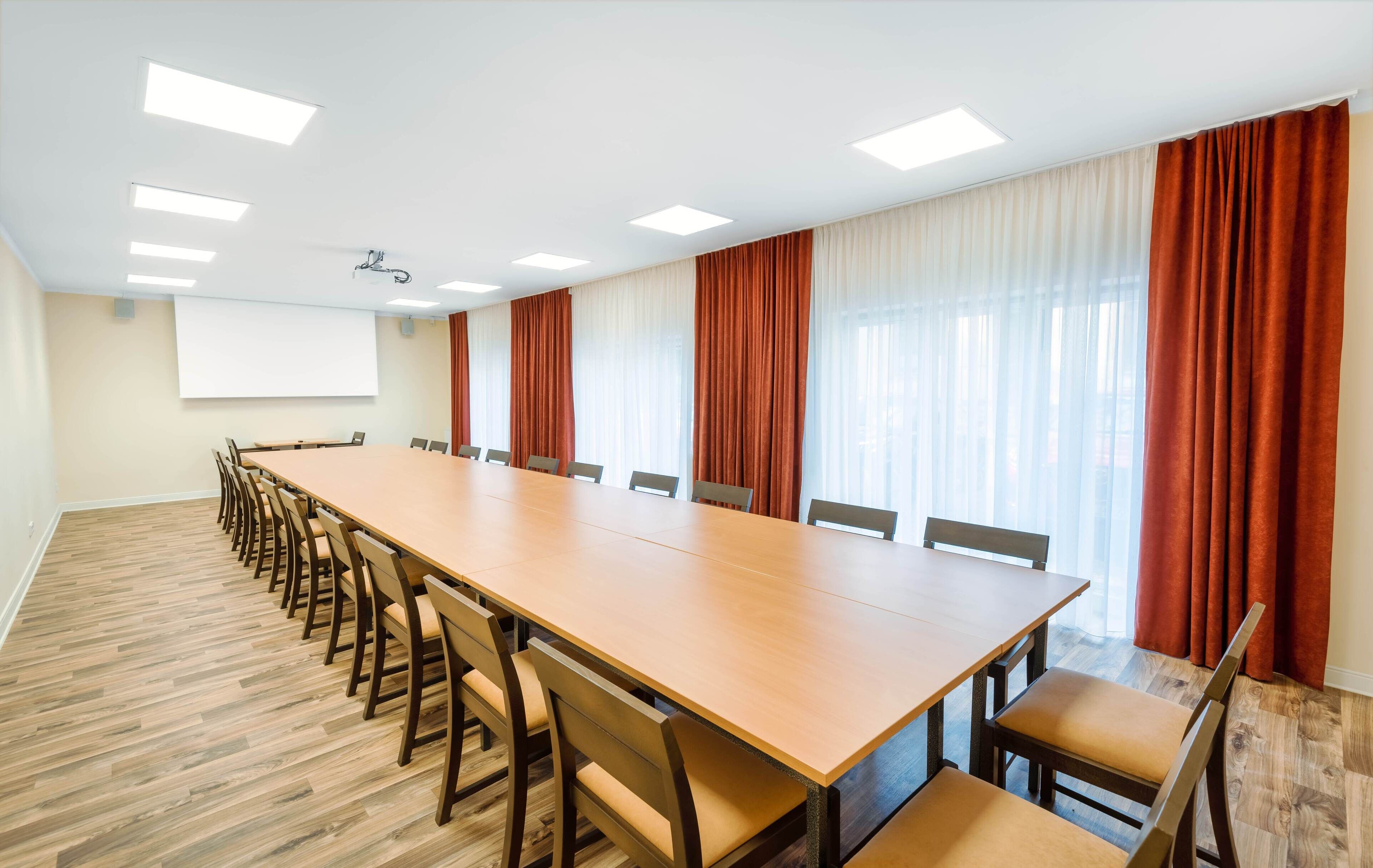 A conference room with a long wooden table, multiple chairs, a projector screen, and large windows with curtains.