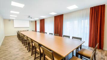 Ein Konferenzraum mit einem langen Holztisch, mehreren Stühlen, einer Projektionsleinwand und großen Fenstern mit Vorhängen.