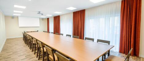 A conference room with a long wooden table, multiple chairs, a projector screen, and large windows with curtains.
