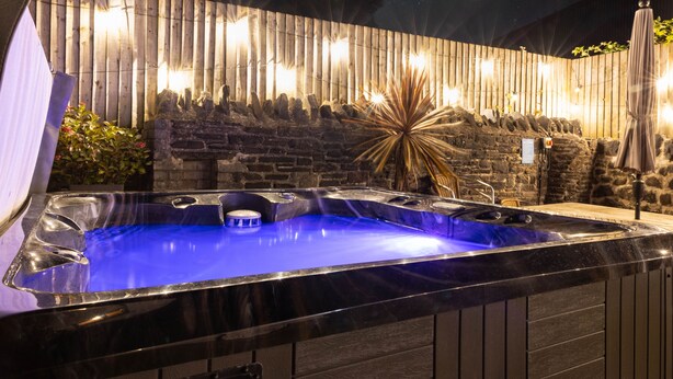 A hot tub with a stone wall backdrop and string lights. - The Coach House 4 bed, 4 bathroom cottage (Porthcawl)
