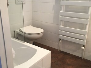 Casa de banho com sanita branca, banheira com divisória de vidro e suporte de toalhas branco.