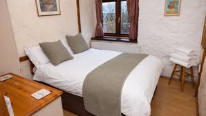 A bedroom with a bed, pillows, a bedside table, a window with curtains, and a small stool.