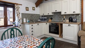 Eine kleine Küche mit weißen Schränken, einer Mikrowelle, einem Herd und einem Spülbecken. Ein Fenster mit Vorhängen und ein Tisch mit gemustertem Tischläufer.