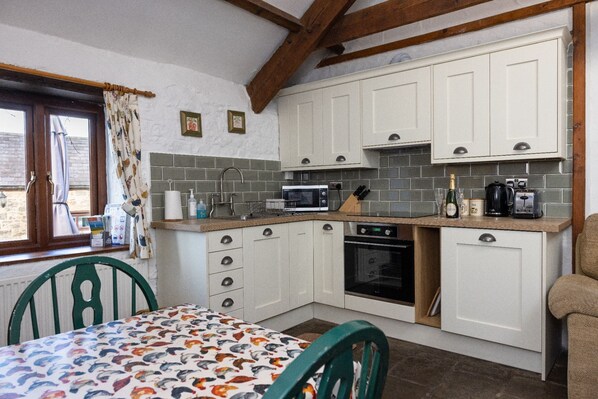 A compact kitchen with white cabinets, a microwave, a stove, and a sink. There is a window with curtains and a table with a patterned tablecloth. - Swallow Cottage - 1 Bedroom - sleeps 3 (Bridgend)