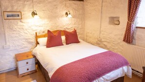 Ein Schlafzimmer mit einem hölzernen Bett, weißen Wänden, einem Nachttisch, zwei Wandlampen und einem Fenster mit gemusterten Vorhängen.