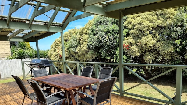 A covered patio with a wooden deck, metal-framed table and chairs, and a grill. - By the Beach (Apollo Bay)