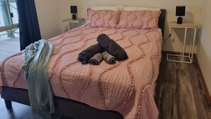 A neatly made bed with a pink quilt and pillows, two rolled-up towels, and a blanket on the left side. There are two bedside tables with lamps, one on each side of the bed. - By the Beach (Apollo Bay)