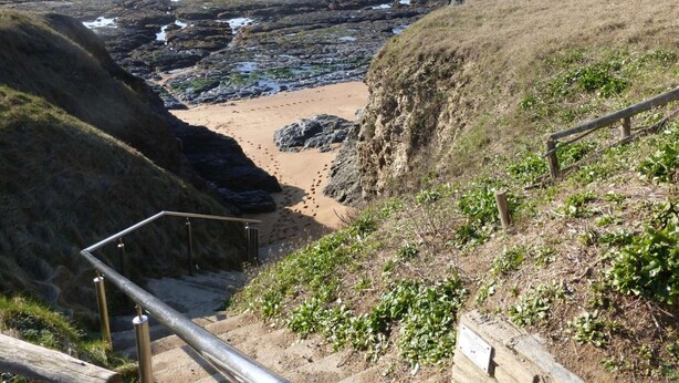 Un sendero costero que lleva a una playa arenosa, con terreno rocoso y una cerca de madera.