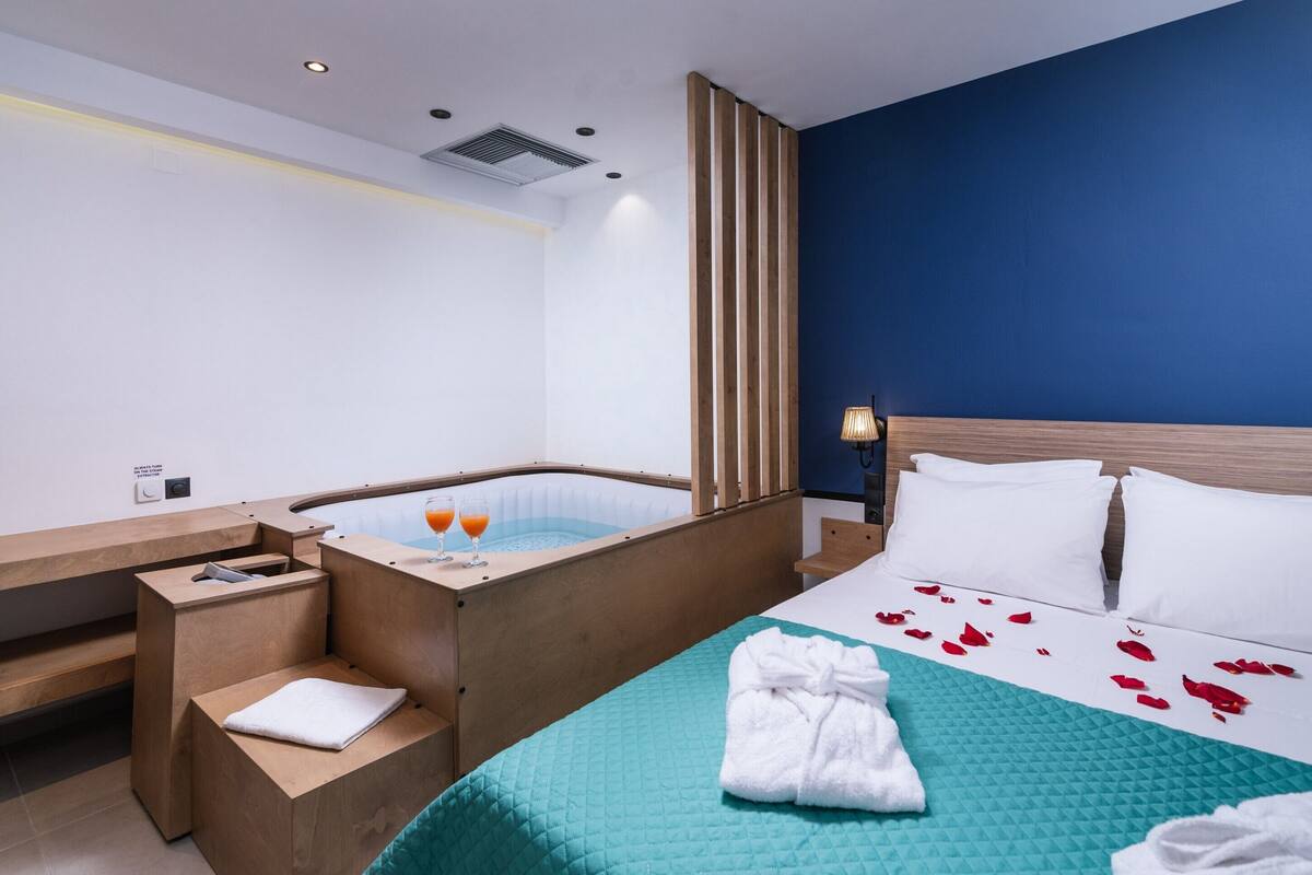 Een moderne hotelkamer met een houten receptiebalie, een badkuip met twee glazen sinaasappelsap, een bed met witte kussens en een turquoise geborduurde spreideken, en een blauwe accentmuur.