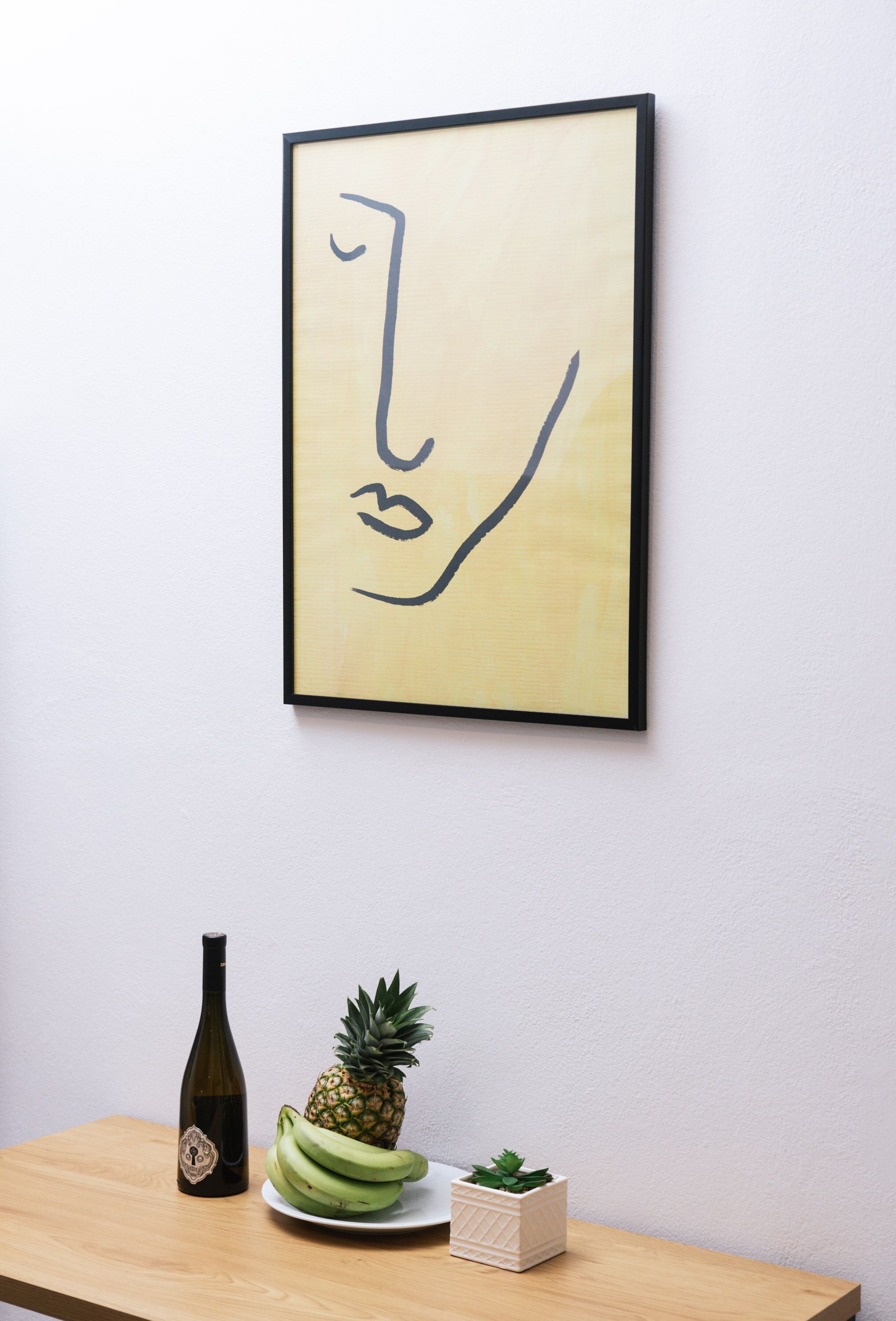 A framed abstract artwork on a wall, a bottle, a pineapple, a plate with bananas, and a small potted plant on a wooden table.