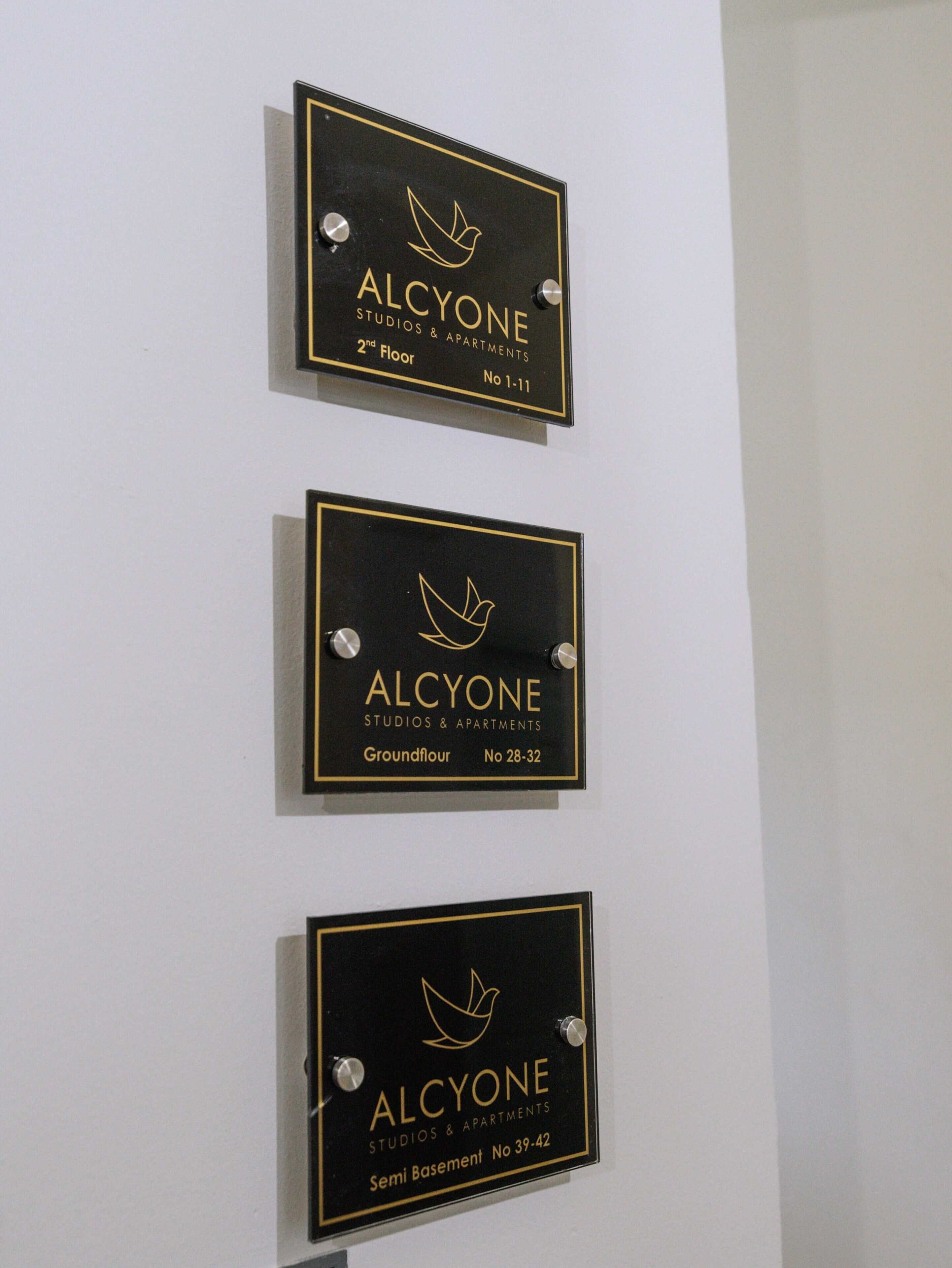 Three black plaques with gold lettering and a bird logo, each indicating a different floor and apartment number for ALCYONE Studios & Apartments.